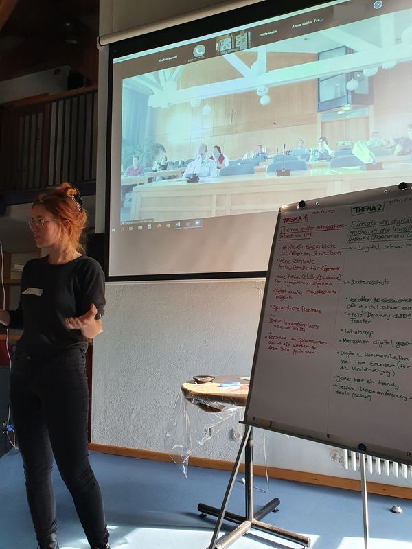 Stehende Frau neben einem Flipchart, im Hintergrund eine Videokonferenz